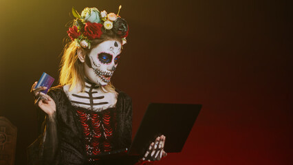 Santa muerte doing online shopping on laptop, paying with credit card in studio. Making electronic...