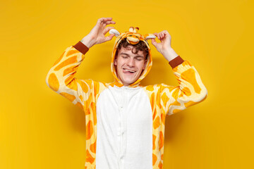 young joyful guy in funny baby giraffe pajamas grimaces and holds his ears on yellow background