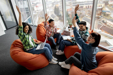 Excited male and female employees putting hands in air after stacking them while working with mobile devices in meeting space. Multiethnic designers in casual outfits motivating team spirit indoors.