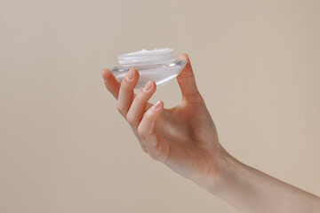 Female hand holding a jar of white moisturizing cream on a beige isolated background. Care for...