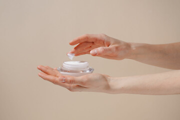 Female hand applying white moisturizing cream from glass jar on finger on beige isolated background. Concept of beauty, cosmetic product, skin treatment and protection