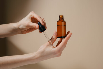 Female hands applying natural mockup serum to skin from brown glass jar on beige isolated...