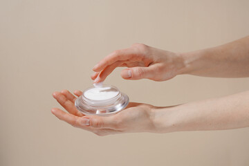 Female hand picking up white moisturizing cream from glass jar with finger on beige isolated background. Concept of cosmetic product, skin protection from weather, beauty brand and treatment