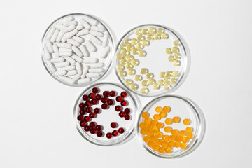 Four laboratory petri dishes with pills capsules in yellow, white, red and orange colors on a white isolated background.