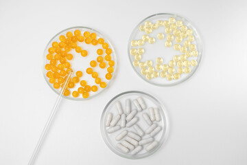 Three petri dishes with orange, yellow and white pill capsules on a white isolated background. Laboratory utensils, dietary supplements and scientific research concept