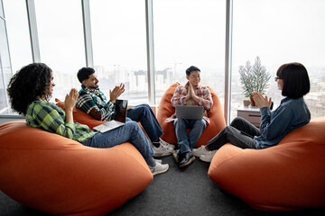 Smiling entrepreneur with laptop on knees holding palms together while team members applauding in office interior. Efficient multiracial employees appreciating workmate for good business results.