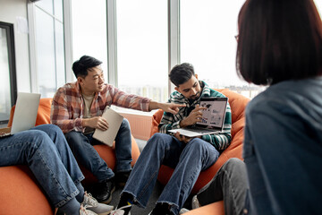 Focus on multicultural men talking about graphic materials on computer while female colleagues sitting on poufs. Business experts analyzing market growth using data visualization in office interior.
