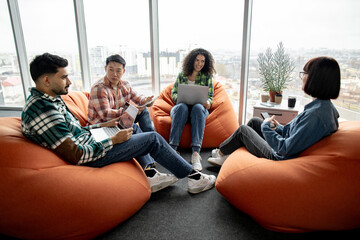 Diverse team of managers in casual wear having conversation while resting on cozy poufs in creative office. Confident collaborators improving corporate strategy using modern technologies in workplace.