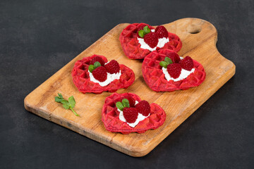 Small Red heart-shaped waffles with cream cheese, raspberries and micro greenery. Waffles with the addition of beet juice. On a serving wooden board. Dark gray background. Valentine's Day