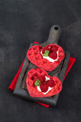 Vegan Red heart-shaped waffles with cream, raspberries and micro greenery. Waffles with the addition of beet juice. On a serving wooden board. Dark gray background.  Valentine's Day