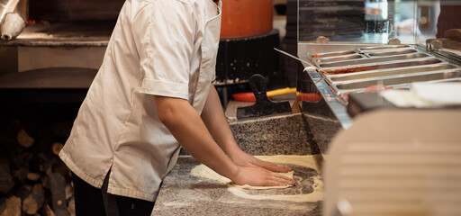 Male chef kneading and rotating dough basis for pizza on the grey marble cooking table in pizzeria's kitchen table