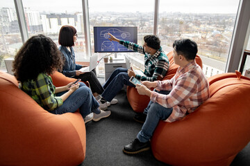 Back view of diverse male and female coworkers examining digital display while staying in open office with terracotta beanbags. Auto designers improving vehicle system using computer programs.