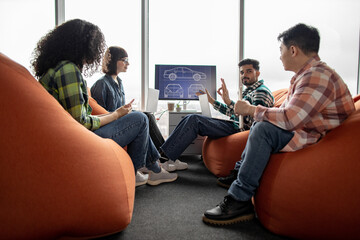 Diverse team of professionals utilizing smart gadgets while being busy at meeting in coworking space with soft poufs. Automotive engineers in casual wear analyzing digital data on display in office.