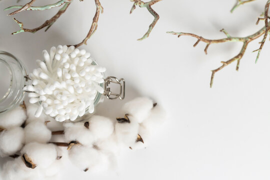 glass jar with cotton ear buds decorated with a dried plant branches and cotton flowers lying on a white background.