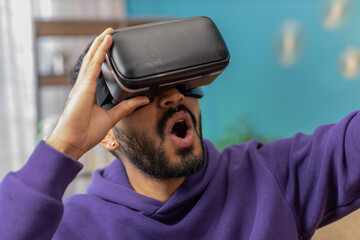 Indian man using virtual reality futuristic technology VR app headset helmet to play simulation 3D 360 video game, watching film movie at modern home apartment. Hindu guy in goggles sitting on couch