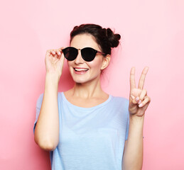 lifestyle, emotion and young people concept: charming smiling young woman show victory sign over pink background
