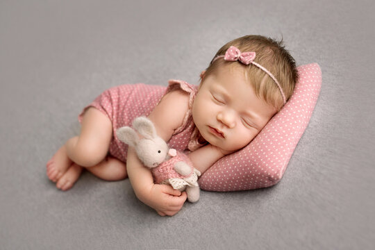 A cute little newborn baby in a pink suit and a pink headband with a butterfly on his head sleeps sweetly. Knitted pear rabbit in hands. Professional macro photo on a grey background.