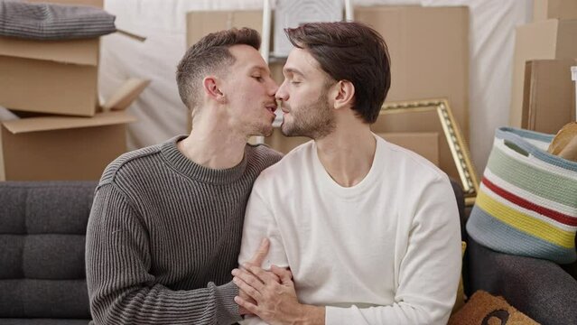 Two men couple sitting on sofa hugging each other kissing at new home