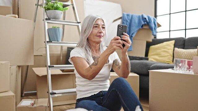 Middle age grey-haired woman recording video by smartphone sitting on floor at new home