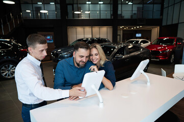 Caucasian married couple chooses a car in a car dealership on digital tablets. 
