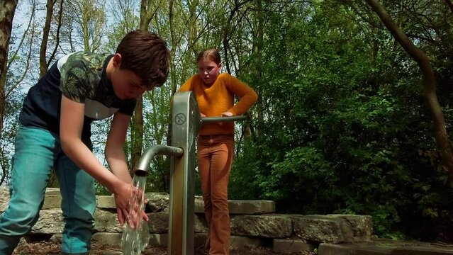 Young children play in a nature playground in the park on sunny spring day. Girl pumps up water with the water pump and boy washes the dirty mud from his hands. Kids outdoor concept, slow motion 