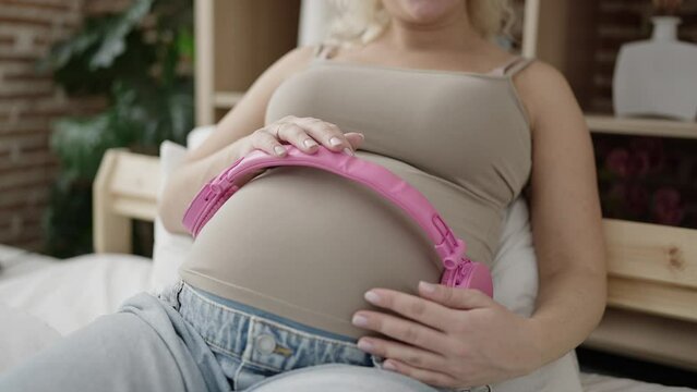Young pregnant woman putting headphones on belly at bedroom