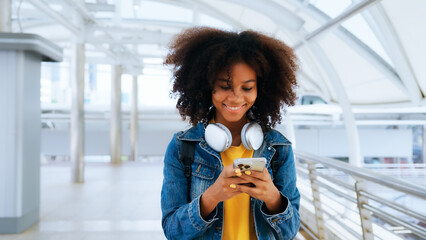 Happy young afro woman listening to playlist music with wireless headphones while wearing yellow shirt and jeans jacket outdoor