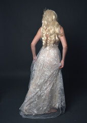 Full length portrait of beautiful women with long blonde hair, wearing fantasy  princess crown and elegant white ball gown, standing pose with hand gesture. Isolated on dark grey studio background.