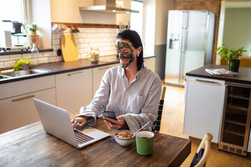 Young adult woman using her phone and laptop while having breakfast in the morning