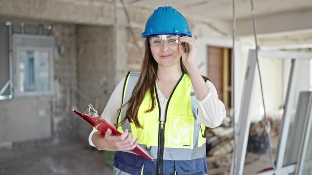 Young beautiful hispanic woman builder smiling confident holding clipboard at construction site