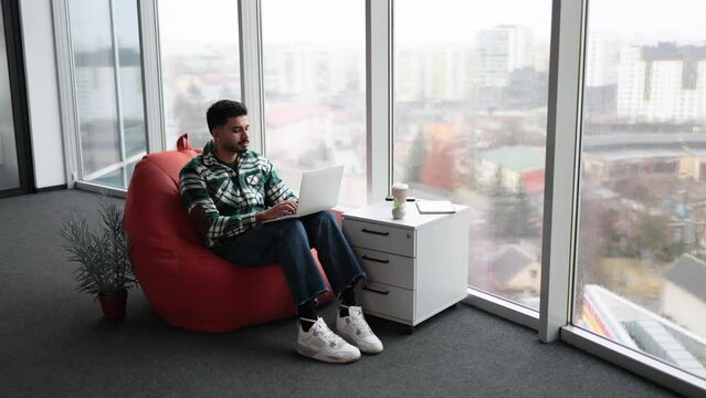 Casually dressed indian programmer working closely on writing and testing codes for new mobile applications in spacious glassy office. Focused man using personal laptop for doing particular job.