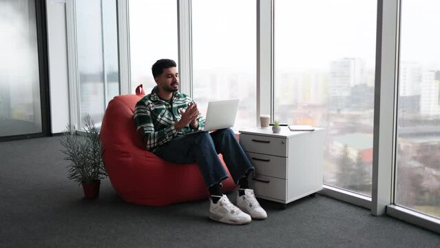 Handsome indian man in casual attire speaking on video call on laptop and gesturing hands in cozy office. Young bearded employee sitting in white spacious workplace with great panoramic view on city.