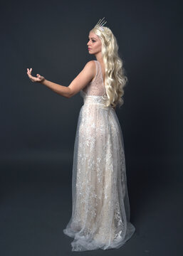 Full length portrait of beautiful women with long blonde hair, wearing fantasy  princess crown and elegant white ball gown, standing pose with hand gesture. Isolated on dark grey studio background.