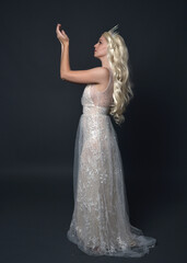 Fototapeta premium Full length portrait of beautiful women with long blonde hair, wearing fantasy princess crown and elegant white ball gown, standing pose with hand gesture. Isolated on dark grey studio background.