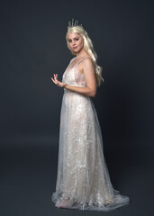 Full length portrait of beautiful women with long blonde hair, wearing fantasy  princess crown and elegant white ball gown, standing pose with hand gesture. Isolated on dark grey studio background. © faestock