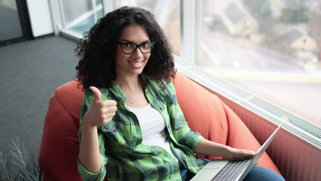 Beautiful multiethnic adult in jeans and T-shirt posing in terracotta beanbag chair with laptop on knees in creative workplace showing sign okay.