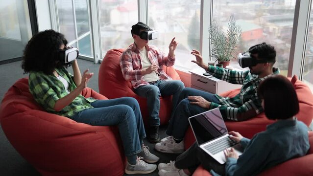 Top view of multiracial teammates working in 3d goggles while young woman in jeans jacket holding portable computer in open office. Focused interior designers making changes to virtual room spaces.