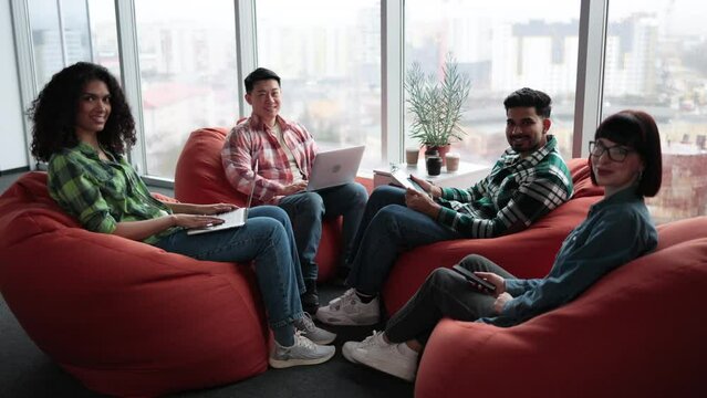 Diverse male and female adults in casual clothes posing on terracotta soft seats with digital gadgets. Multiethnic collaborators planning presentation for colleagues in shared workspace.