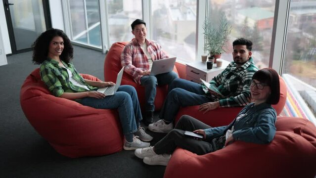 Top view of diverse male and female adults in casual clothes posing on terracotta soft seats with digital gadgets. Multiethnic collaborators planning presentation for colleagues in shared workspace.