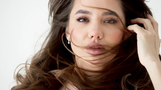 Smiling sophisticated young woman with dark hair blowing in the wind looks at the camera. Happy healthy cute woman beauty model advertising skin or hair care product in slow motion.