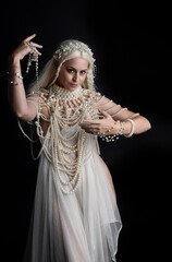 Close up portrait of beautiful blonde female model wearing an extravagant pearl jewelled gown. Gestural arms reaching out,  isolated on a black studio background.