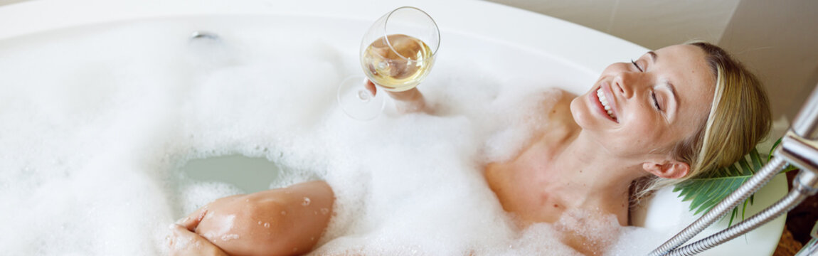 Top view on happy joyful young woman lying in bubbles in bathtub with glass of white wine.