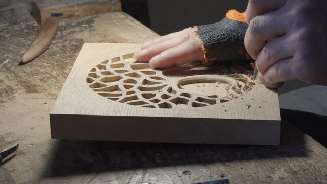 Close up hands of a wood carver using a small chisel to make a relief carving of a tree in circle shape from a flat square piece of wood