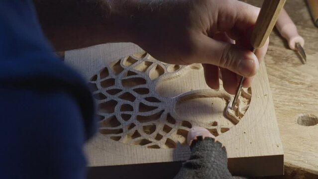 Close up hands of a wood carver using a small chisel to make a relief carving of a tree in circle shape from a flat square piece of wood, shot in slow motion