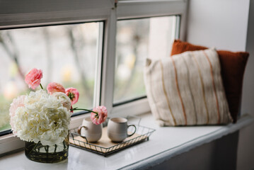 Window white sill with flowers, cups of coffee, cozy pillows, space for decoration. Interior of home room, house design. Modern decor for indoor place.  Comfortable apartment. Spring still life scene.