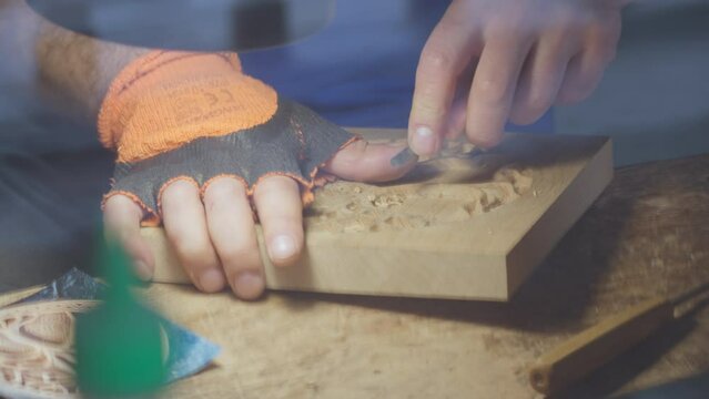 Close up hands of a wood carver using a small chisel to make a relief carving of a tree in circle shape from a flat square piece of wood, shot through a glass window showing outdoor reflection