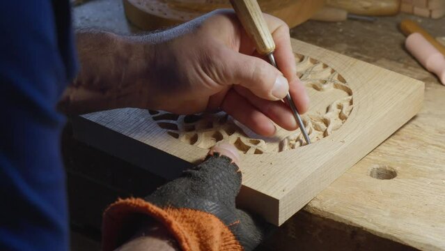 Close up hands of a wood carver using a small chisel to make a relief carving of a tree in circle shape from a flat square piece of wood, shot through a glass window showing outdoor reflection