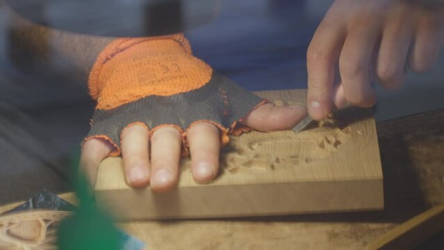 Close up hands of a wood carver using a small chisel to make a relief carving of a tree in circle shape from a flat square piece of wood, shot through a glass window showing outdoor reflection