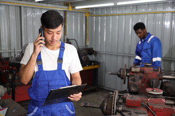 Multiethnic technicain mechanic using metal lathe machine operate polishing car disc brake with smart mobile phone and clipboard at garage. Maintenance automotive and inspecting vehicle part concept