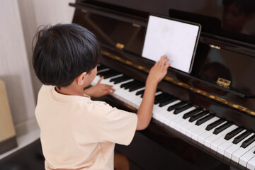 Kid asian black hair boy sitting and playing piano with tablet in living room house indoor. Musical and relaxation makes them happiness. Health care lifestyle concept.